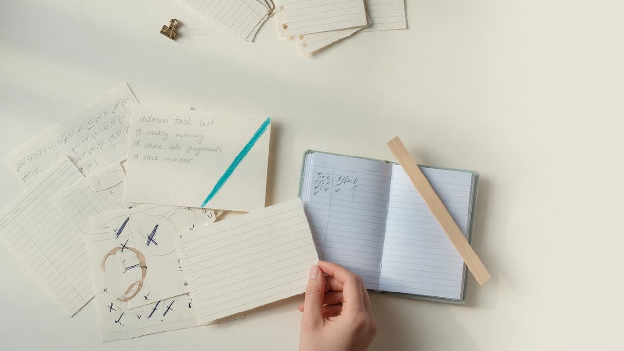 A small-business desk with workflow notes, a notebook and a teal pencil marking one recurring administrative task for review